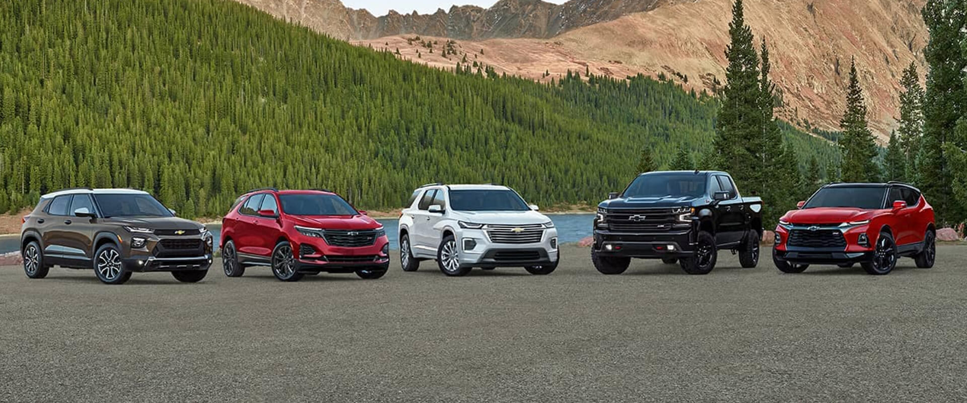 Lineup of Chevrolet vehicles including SUVs and a pickup truck parked by a scenic lake with forested mountains in the background, showcasing variety and versatility.